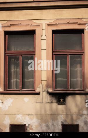 Two tall vintage wooden windows set in a weathered beige facade with peeling plaster and decorative architectural trim Stock Photo