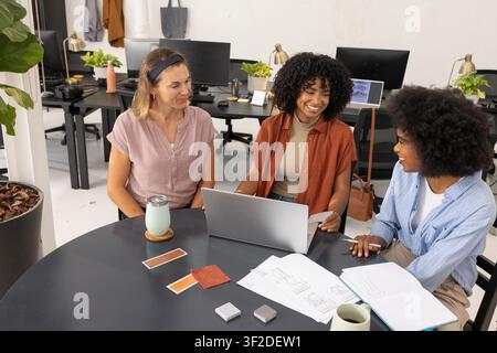 Diverse female coworkers collaborating, reviewing laptop, plans and swatches in open office. Teamwork, design, materials, textiles, diagrams, discussion, coworking Stock Photo