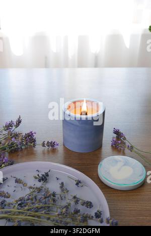 Scented soy candle burning in blue marble concrete holder on wooden dining table near ceramic tray with dried lavender flowers for cozy morning Stock Photo