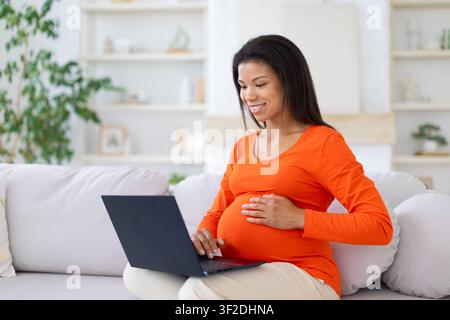 Smiling Black pregnant woman working on laptop, sitting on sofa at home Stock Photo