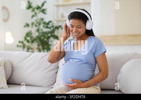 Smiling Black pregnant woman listening music through wireless headphones while holding belly Stock Photo