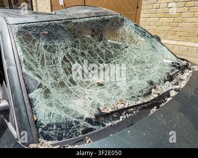Damaged car windshield showing shattered glass debris, web-like pattern of cracks, dark colored car parked in front of light brick wall, natural dayli Stock Photo