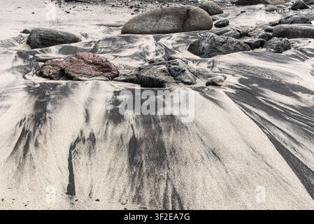 Dark patches forming natural designs in the light beach sand, flanked by scattered stones, creating an abstract visual. Stock Photo