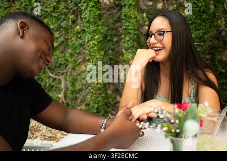 Multicultural Venezuelan and Dominican couple laughing and holding hands at an outdoor table, enjoying a casual date with drinks and flowers in a gree Stock Photo