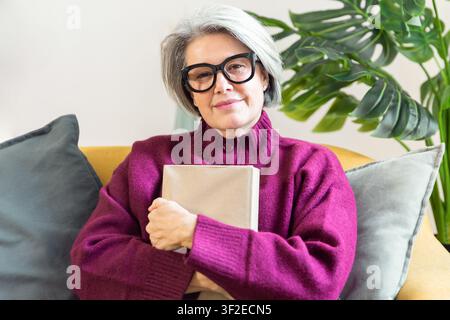 Senior woman with gray hair and glasses sits on a sofa holding a book, enjoying a calm home routine focused on wellness, comfort, and mindful relaxati Stock Photo