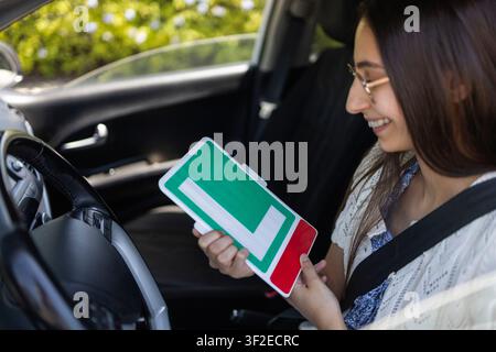 Smiling young woman wearing a seat belt in a parked car, holding a learner L-plate sign and looking ahead, symbolizing driving lessons, licensing, and Stock Photo