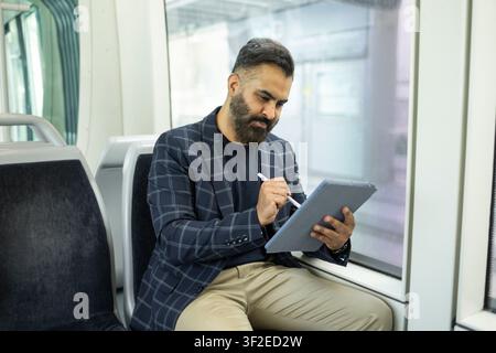 Indian businessman in a check blazer sits on a train by the window, using a tablet and stylus for work, planning tasks during an urban commute. Stock Photo