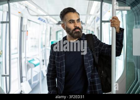 Bearded Indian man commuting on a modern train, holding a pole with a backpack. Urban public transport and daily travel concept with bright interior. Stock Photo