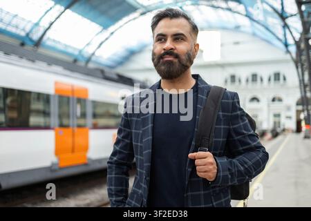 Confident Indian businessman with beard and backpack walking on a modern train platform, wearing a checked blazer, commuting for work travel in an urb Stock Photo
