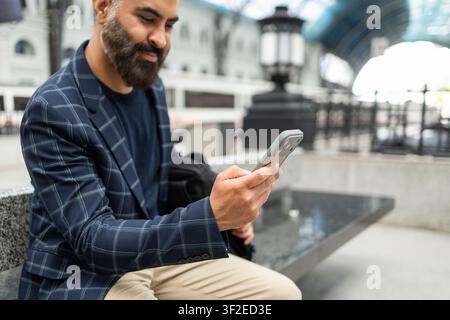 Indian businessman in a checked blazer sits at a modern train station, smiling while using a smartphone, suggesting commuting, travel planning, and mo Stock Photo