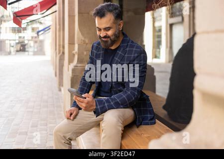 Indian businessman in a plaid blazer sits on a wooden bench in an urban arcade, checking his smartphone, relaxed and focused during a city break. Stock Photo