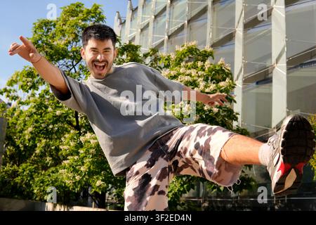 Cheerful fashionable Hispanic man in casual streetwear jumps midair outdoors, pointing and smiling, with modern glass building and green trees in the Stock Photo
