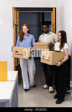 Three coworkers carry cardboard boxes through a doorway during an office move, symbolizing relocation, teamwork, and a fresh start in a modern workpla Stock Photo