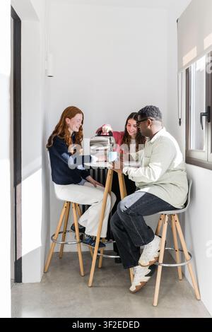 Three friends sit on stools around a small kitchen table, pouring coffee from a machine and chatting in a bright, modern office nook with sunlight and Stock Photo
