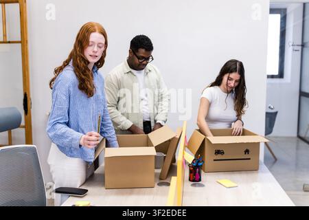 Diverse coworkers pack cardboard boxes at a modern office table, organizing supplies and stationery for a workplace move, relocation, or new workspace Stock Photo
