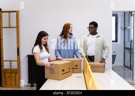 Three young coworkers pack and organize cardboard boxes on a table in a modern office, preparing for relocation, shipping, or workspace setup with tea Stock Photo