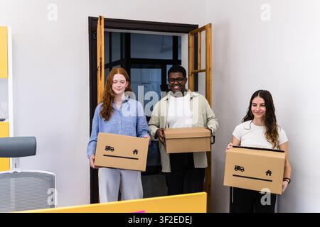 Three employees carry cardboard boxes through a doorway in a modern office, suggesting relocation, new workspace setup, teamwork, and a fresh start in Stock Photo