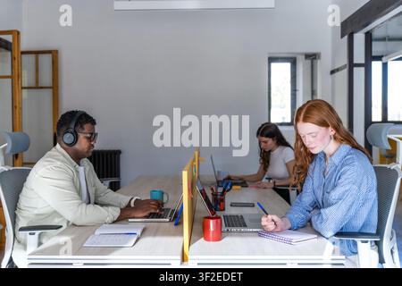 Diverse coworkers work quietly at a long shared desk in a bright open office. A man with headphones types on a laptop while a red-haired woman takes n Stock Photo