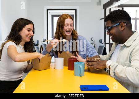 Three coworkers share a casual takeout lunch at a bright office table, using chopsticks and coffee mugs while laughing together during a relaxed break Stock Photo
