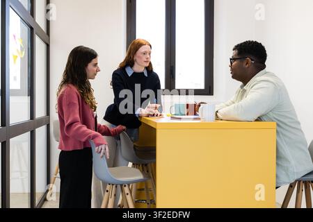 Three coworkers meet around a yellow counter in a bright modern office, sharing ideas and notes during a casual team discussion with coffee mugs and l Stock Photo