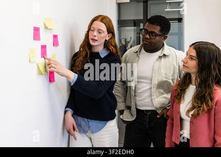 Diverse coworkers collaborate in a modern office, reviewing colorful sticky notes on a whiteboard during an agile planning session, discussing tasks, Stock Photo