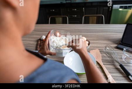 Overhead view of a woman mixing flour and eggs in a bowl on a kitchen counter, preparing handmade pasta dough with whisk and wooden spatula for a home Stock Photo