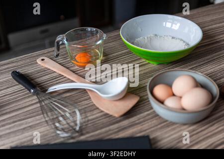Baking setup on a kitchen table with eggs in a bowl, a measuring cup holding an egg yolk, flour in a mixing bowl, and a whisk with wooden spatula and Stock Photo
