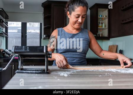 Woman cooking in a modern home kitchen, rolling fresh handmade pasta dough on a floured counter beside a pasta machine, preparing an Italian-style mea Stock Photo
