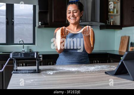 Smiling woman in a home kitchen stretches fresh handmade pasta dough over floured hands beside a pasta machine, preparing ingredients for an authentic Stock Photo