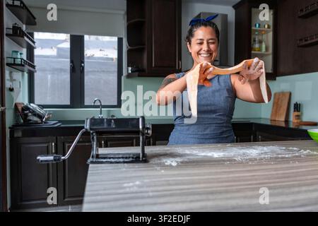 Smiling woman stretches fresh handmade pasta dough in a modern home kitchen, preparing noodles on a floured counter with a pasta machine, enjoying hom Stock Photo