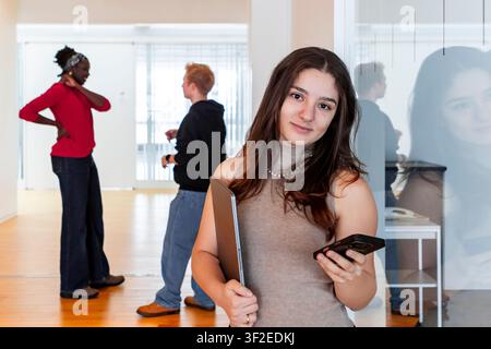 Female college student stands in a bright classroom holding a laptop and smartphone, smiling at the camera while classmates discuss in the background. Stock Photo