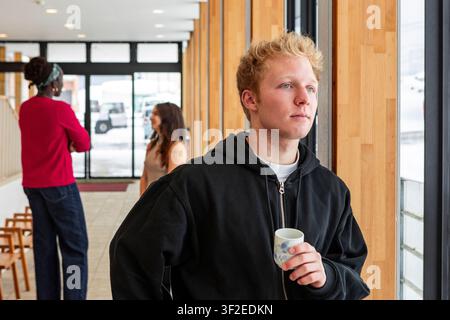 Young student holding a cup of coffee stands by a large window in a campus hallway, with other multiethnic students in the background on a bright wint Stock Photo