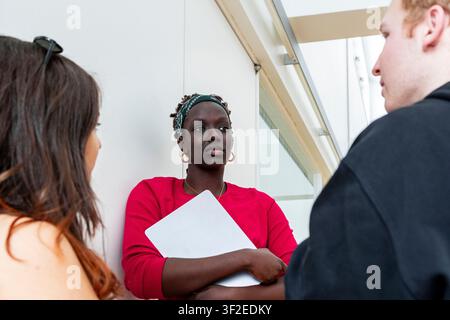 Multiethnic student group meets in a bright campus hallway, holding notebooks and papers while discussing coursework, teamwork, and academic planning Stock Photo
