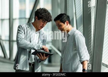 Two business colleagues stand in a bright modern office corridor, reviewing charts on a tablet together, discussing strategy, teamwork, and a digital Stock Photo