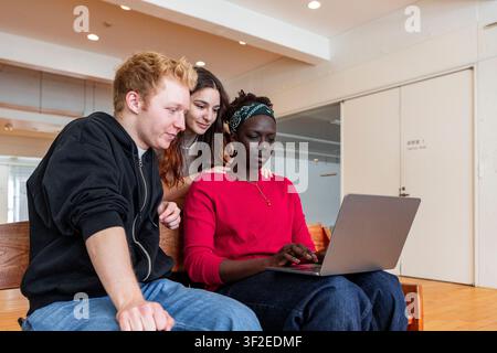 Diverse college students work together on a laptop in a bright campus lounge, sharing ideas for a project, studying as a team, and supporting digital Stock Photo
