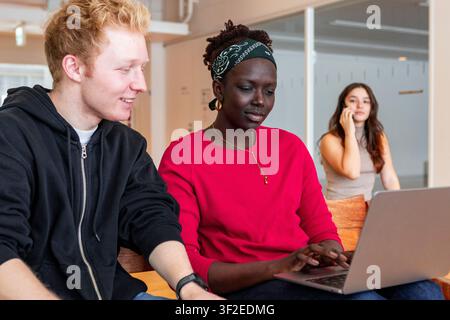Multiethnic college students collaborate at a table, one focused on a laptop while another talks on a smartphone in the background in a bright modern Stock Photo