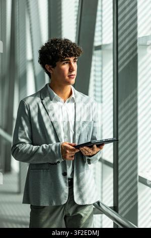 Young businessman in a light suit holding a tablet, standing by a sunlit office window with shadow patterns, looking away thoughtfully in a modern cor Stock Photo