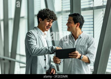 Two male coworkers collaborate in a bright modern office, reviewing charts on a tablet and discussing project updates, teamwork, strategy, and digital Stock Photo