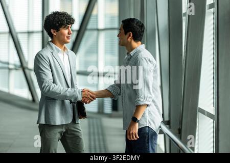 Two business partners shake hands in a bright, modern office corridor with glass walls, sealing an agreement during a professional meeting and network Stock Photo
