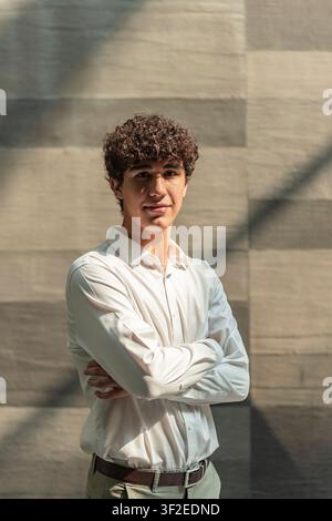 Confident young professional in a white shirt stands with arms crossed by a textured wall, lit by natural sunlight and soft shadows, ideal for busines Stock Photo