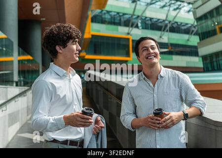 Two young business colleagues stand in a bright modern office atrium, holding takeaway coffee and chatting casually, suggesting teamwork, networking, Stock Photo