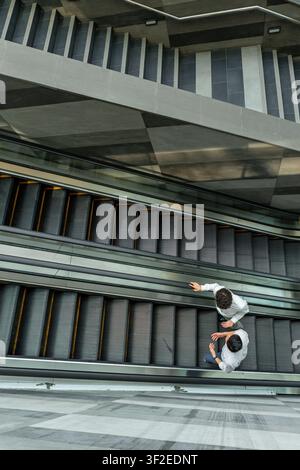 Top view of two people riding parallel escalators inside a modern concrete and glass building, creating strong diagonal lines and an abstract urban tr Stock Photo