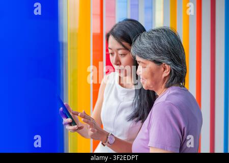 Senior Asian mother and adult daughter stand together using a smartphone, sharing technology tips and support, with a colorful wall background. Stock Photo