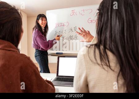 Smiling teacher leads an adult Japanese lesson in a small classroom, writing hiragana and greetings on a whiteboard as students follow along with a la Stock Photo