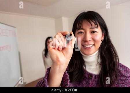 Smiling Japanese language teacher holding marker and looking at camera during an adult Japanese lesson in classroom, with whiteboard in background. Stock Photo