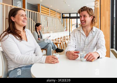 A diverse group of students relax on campus during a break. They gather around a table, sharing coffee, laughter, and conversation in a bright, modern Stock Photo