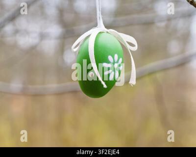 Decorative green painted Easter egg with white flower pattern hanging on a tree branch outdoors. Stock Photo