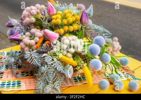 Decorative spring bouquet featuring yellow craspedia, pink tulips, blue thistle, fuzzy stems, foliage on bright yellow table with woven rustic textile Stock Photo