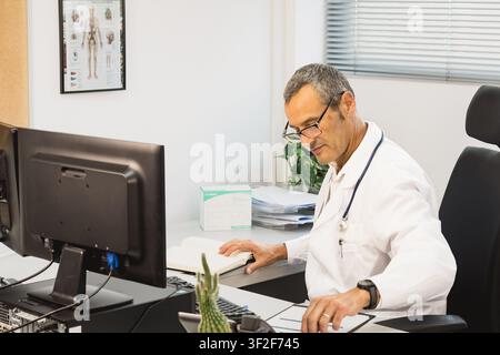 Male doctor seated at his desk in a medical office, reviewing a notebook with glasses on, beside a computer monitor in a bright clinical setting. Stock Photo