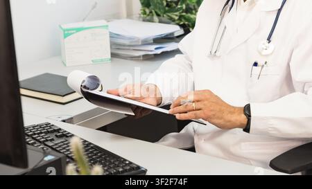 Close up of a doctor holding and reviewing paperwork at his desk, with keyboard, monitor and clinical office details visible. Stock Photo
